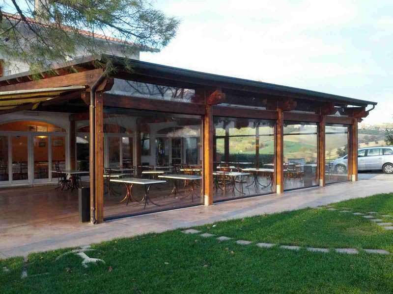 Veranda in legno di un ristorante chiusa su tutti i lati da teli trasparenti, con i tavoli all'interno e le colline sullo sfondo