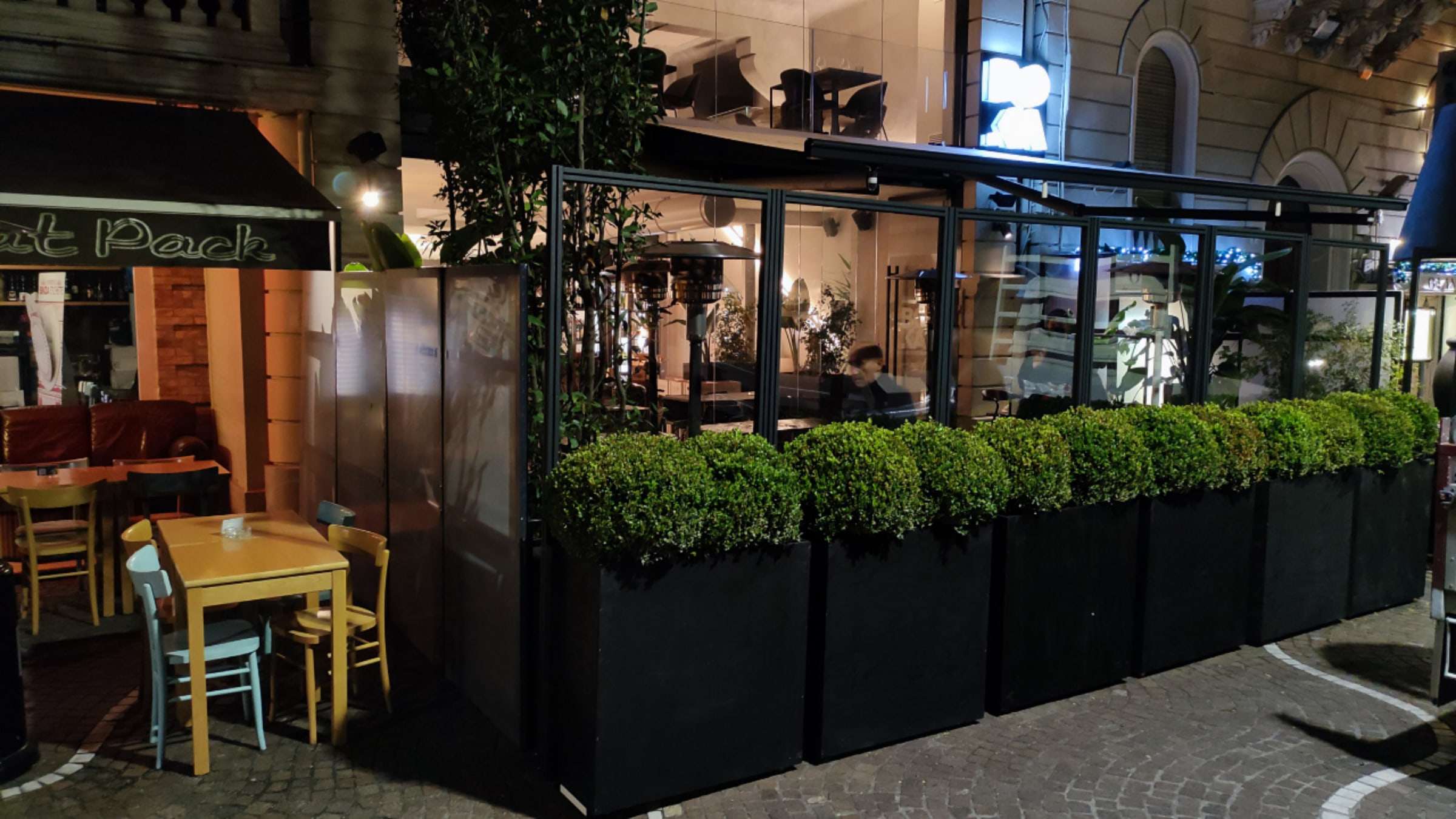 Dehors di un bar la sera, chiuso da vetrate con fioriere e luci calde