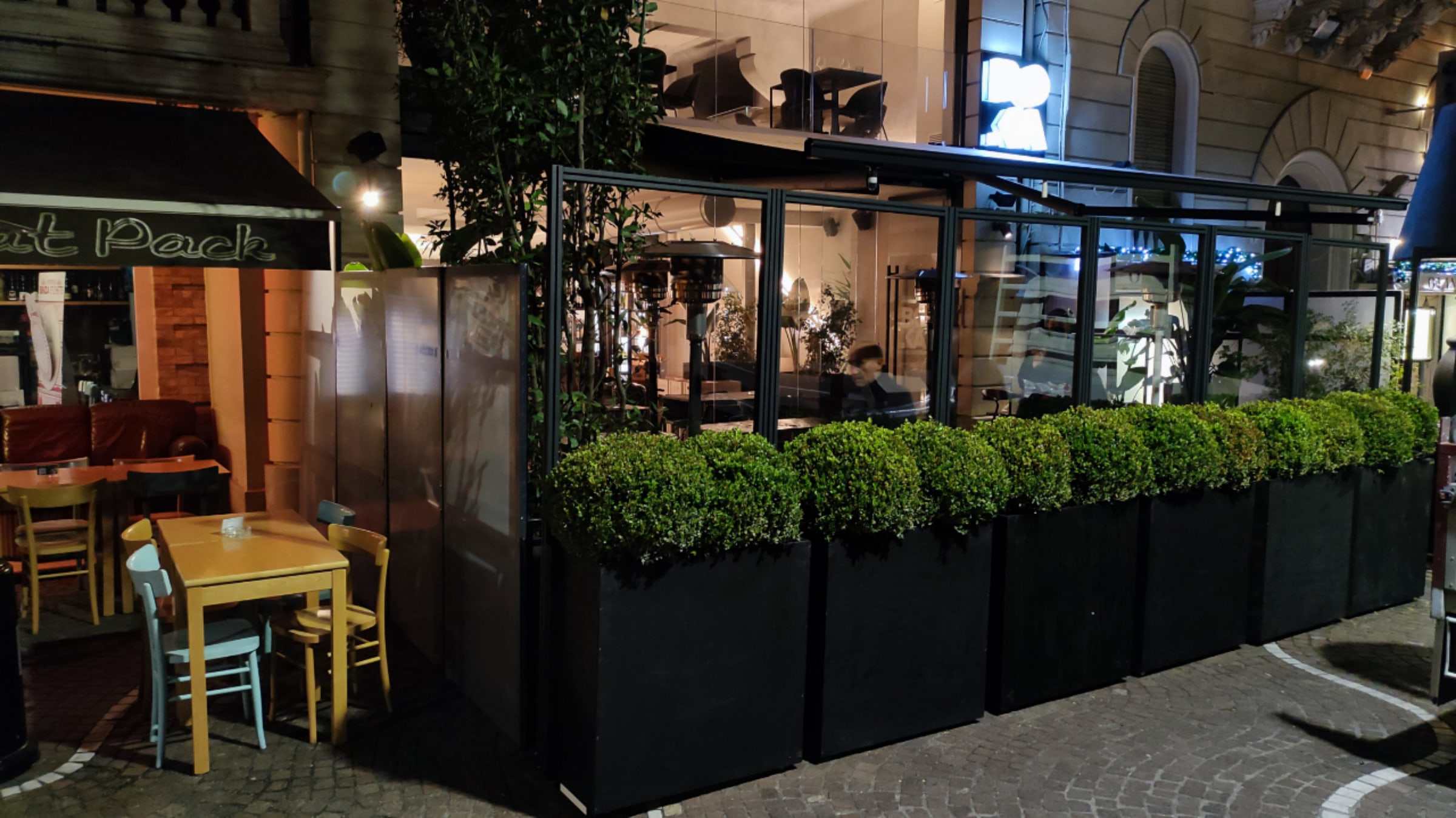 Dehors di un bar la sera, chiuso da vetrate con fioriere e luci calde