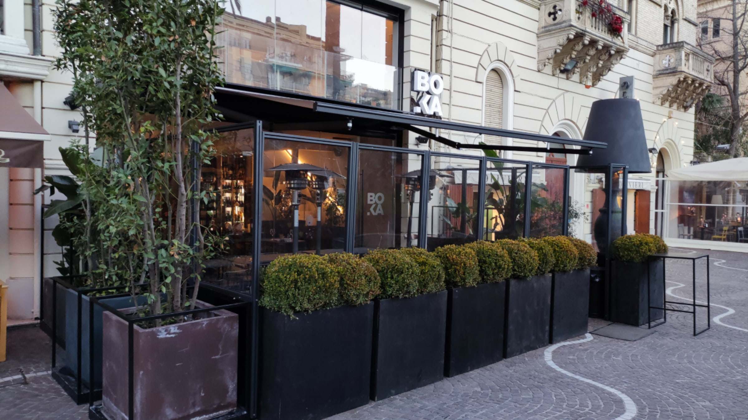 Dehors serale di un bar con struttura scura, chiusure trasparenti e fioriere davanti a un palazzo storico