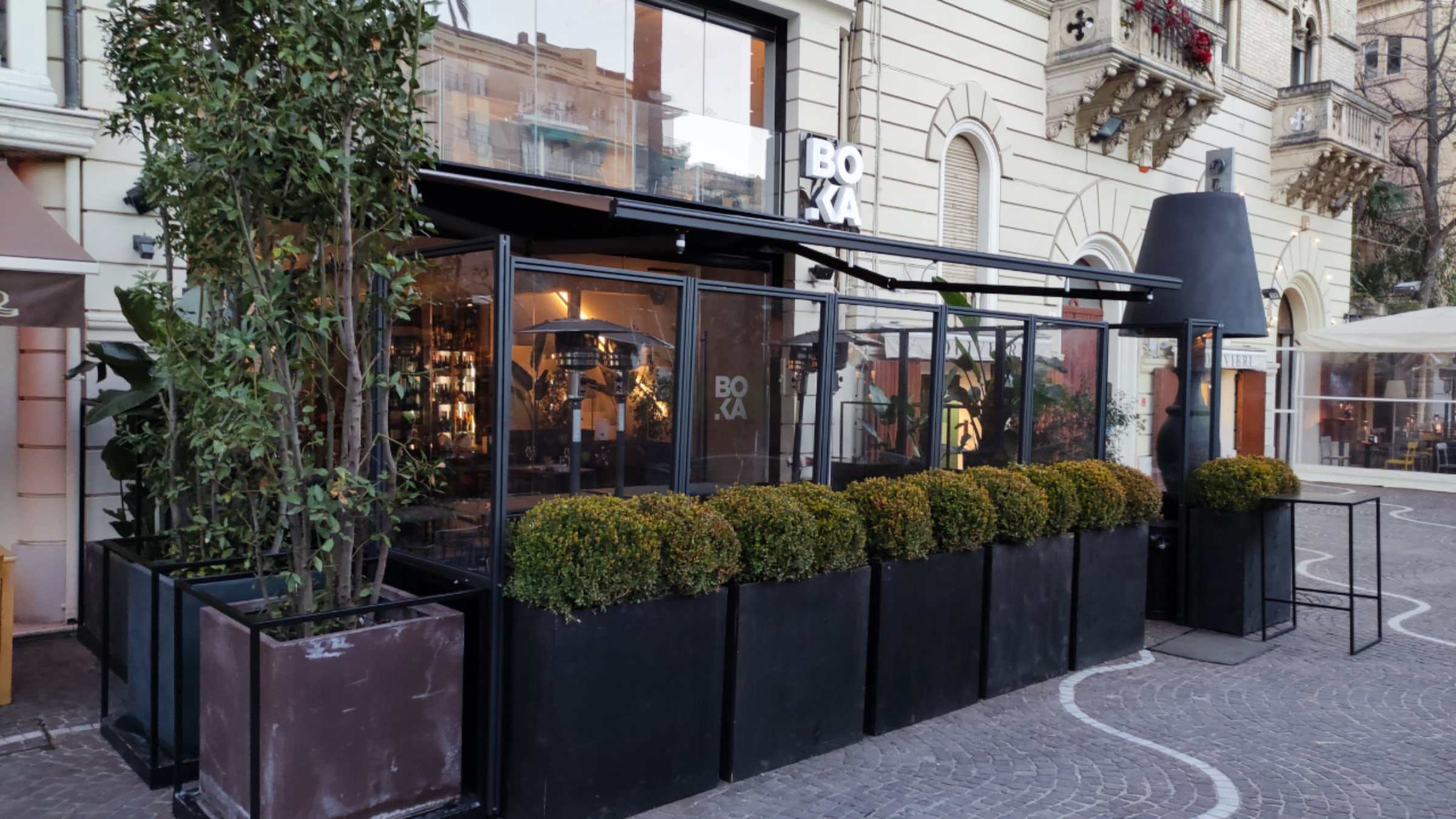 Dehors serale di un bar con struttura scura, chiusure trasparenti e fioriere davanti a un palazzo storico