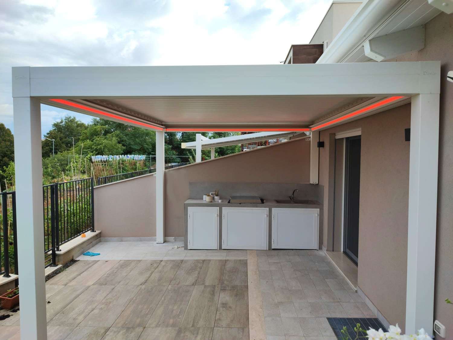 Pergola bioclimatica bianca con LED integrati nel telaio sopra una cucina da esterno