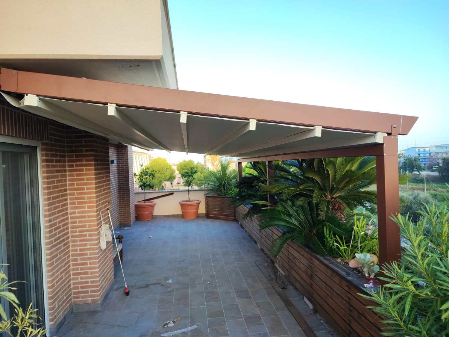 Pergotenda marrone con telo chiaro su terrazzo con fioriere