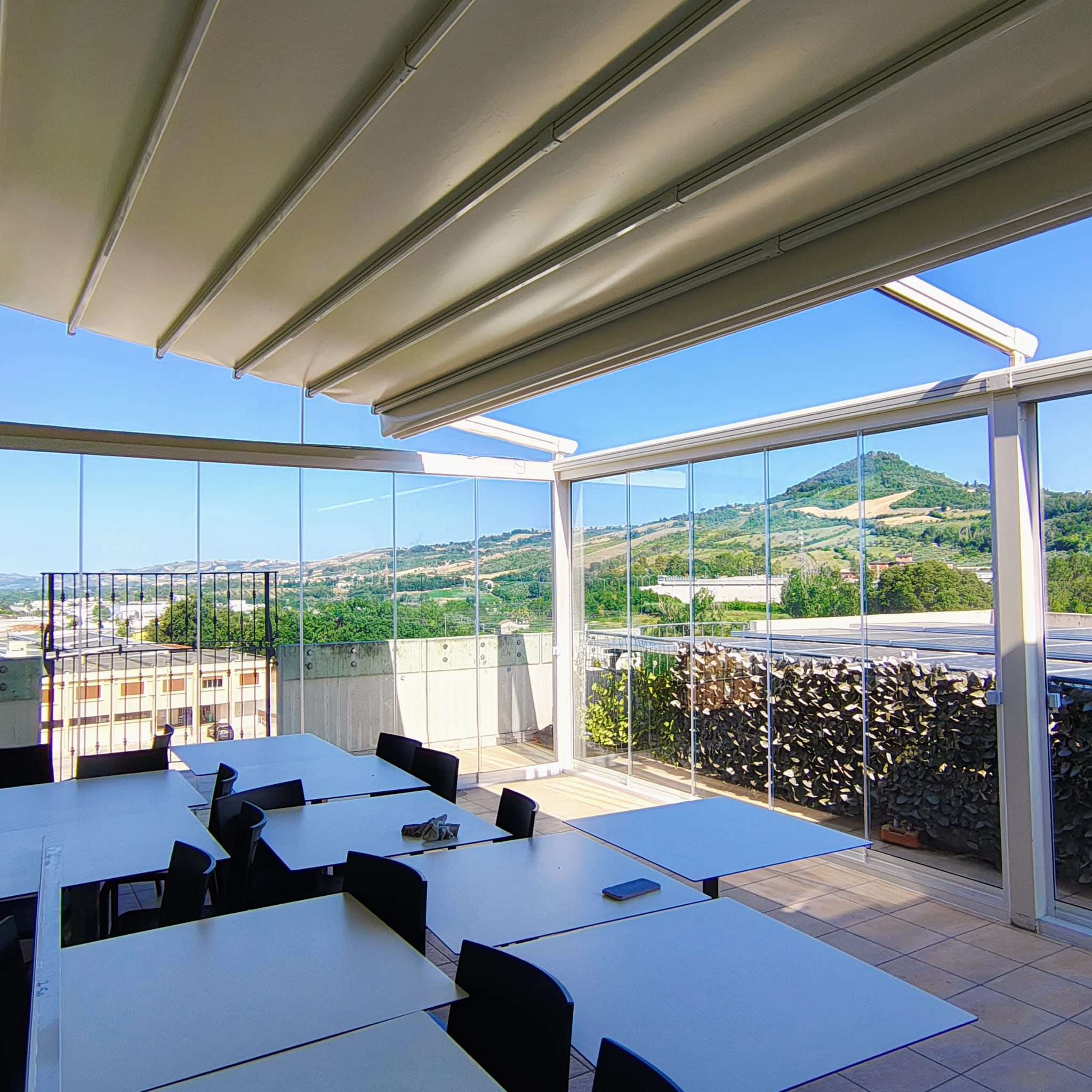 Terrazza di un ristorante coperta da pergotenda e chiusa da vetrate con vista sulle colline