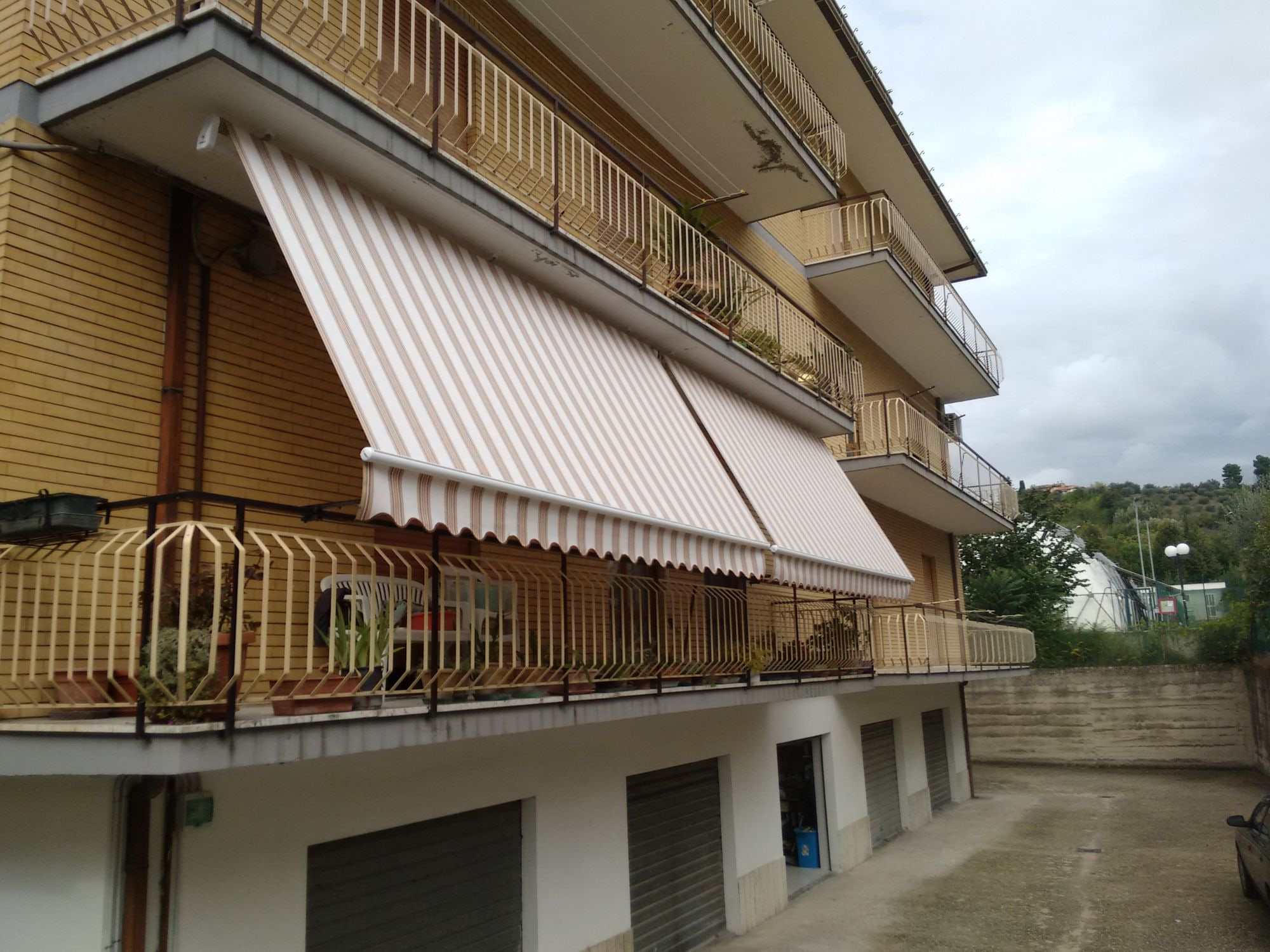 Due tende a caduta con braccetti a righe beige e bianche sul balcone di una palazzina in mattoni