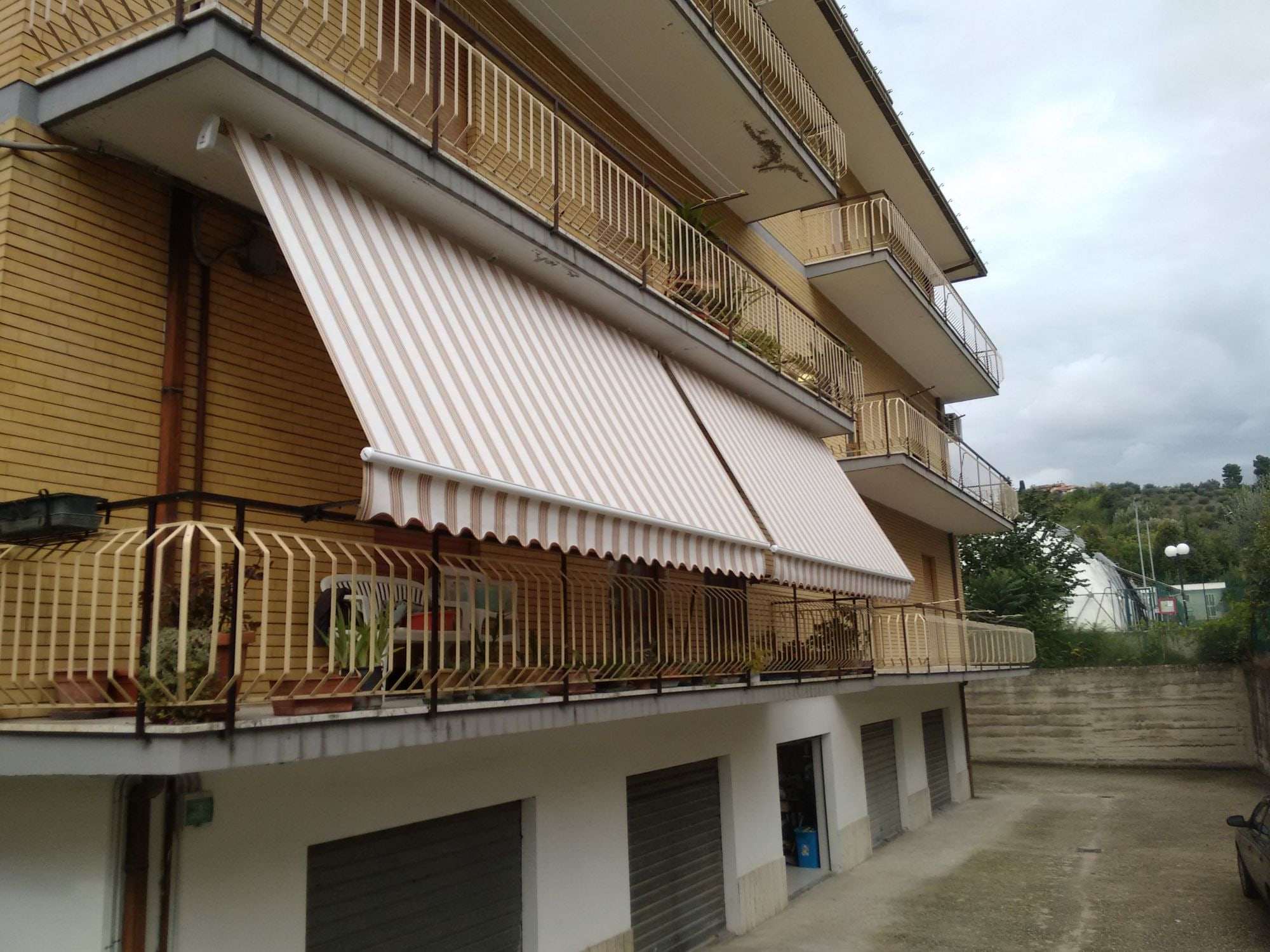 Due tende a caduta con braccetti a righe beige e bianche sul balcone di una palazzina in mattoni