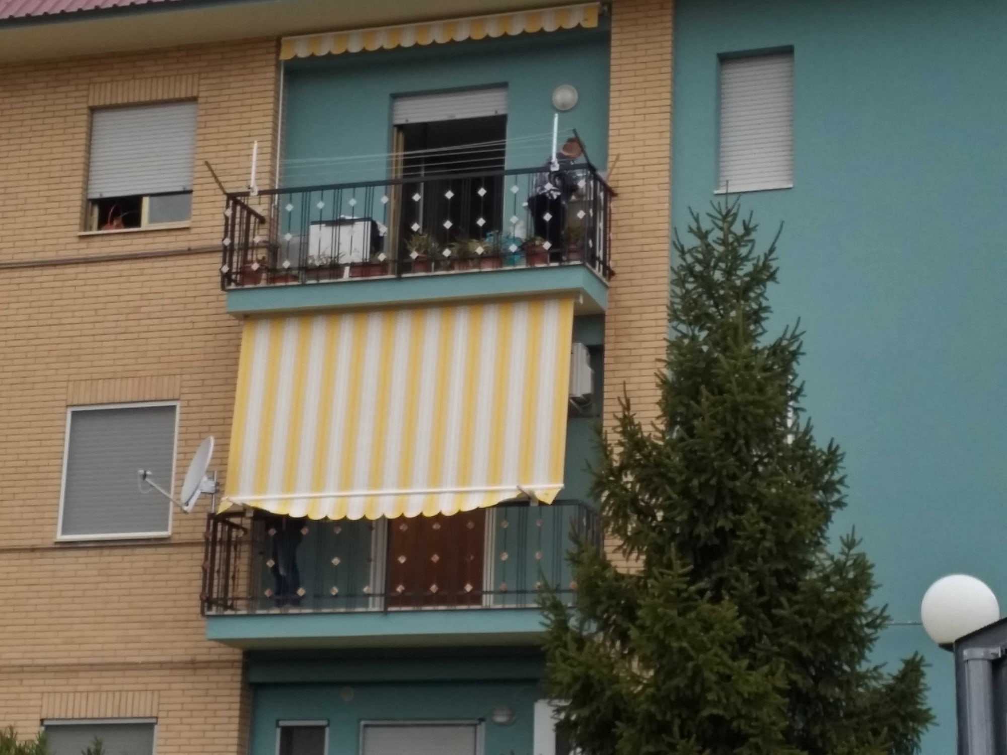 Tenda a caduta con braccetti a righe gialle e bianche su un balcone di una palazzina color mattone e verde acqua