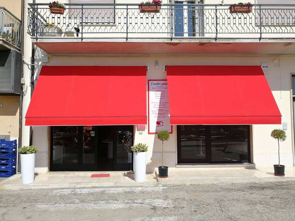 Due tende a caduta con telo rosso sopra le vetrine di un negozio, sotto un balcone